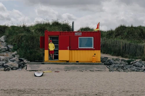 A red and yellow beach hut on the sand at Portmarnock, Dublin, a single figure standing beside it under a cloudy sky, marram fencing along the dunes behind.