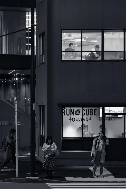 A lit corner shopfront on a dark street at night, figures passing below, Tokyo, in black and white.