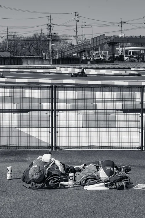 Black and white view through a trackside safety fence at a motor racing circuit, with a person resting on the ground beside bags in the foreground