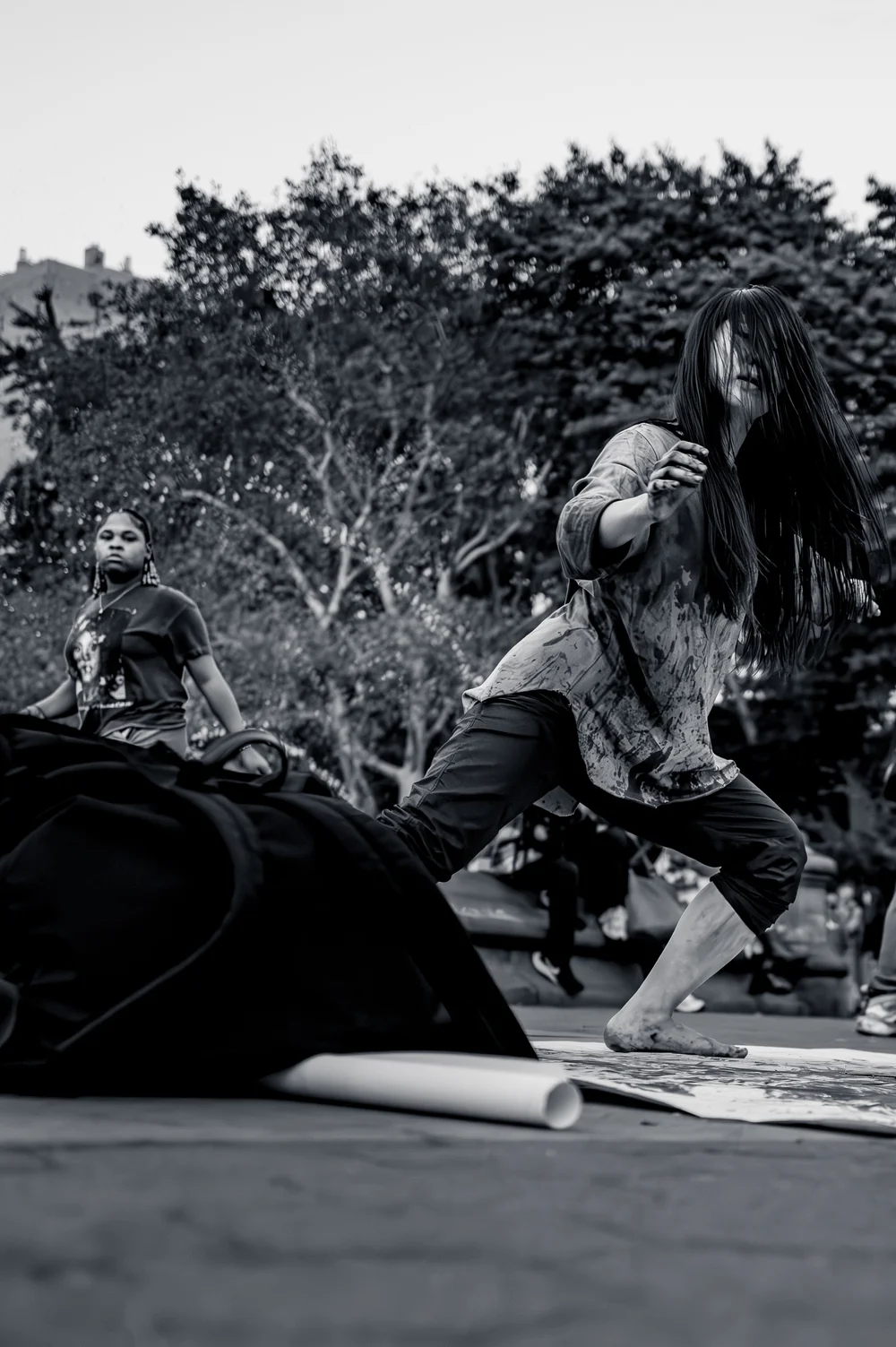 A dancer with long dark hair caught mid-movement low to the ground, trees behind her in Washington Square Park, New York, in black and white.