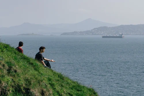 Two people sitting on the grassy cliff at Howth Head, Dublin, looking out over the bay toward distant mountains, a ship on the horizon.