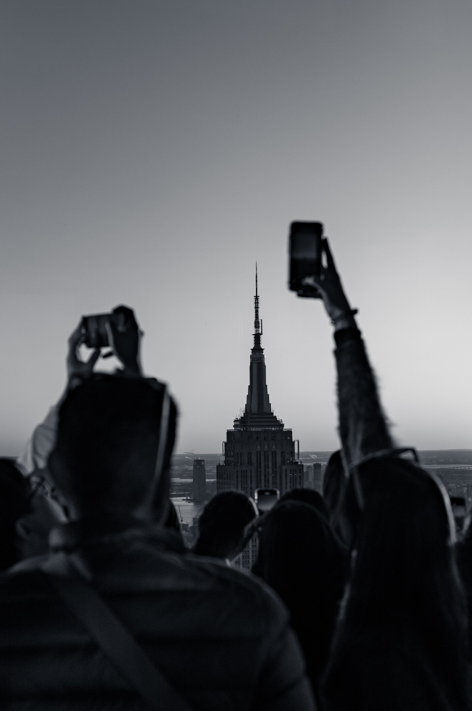 A crowd seen from behind with phones raised at Summit One Vanderbilt, the spire of the Empire State Building centred between them, New York, in black and white.