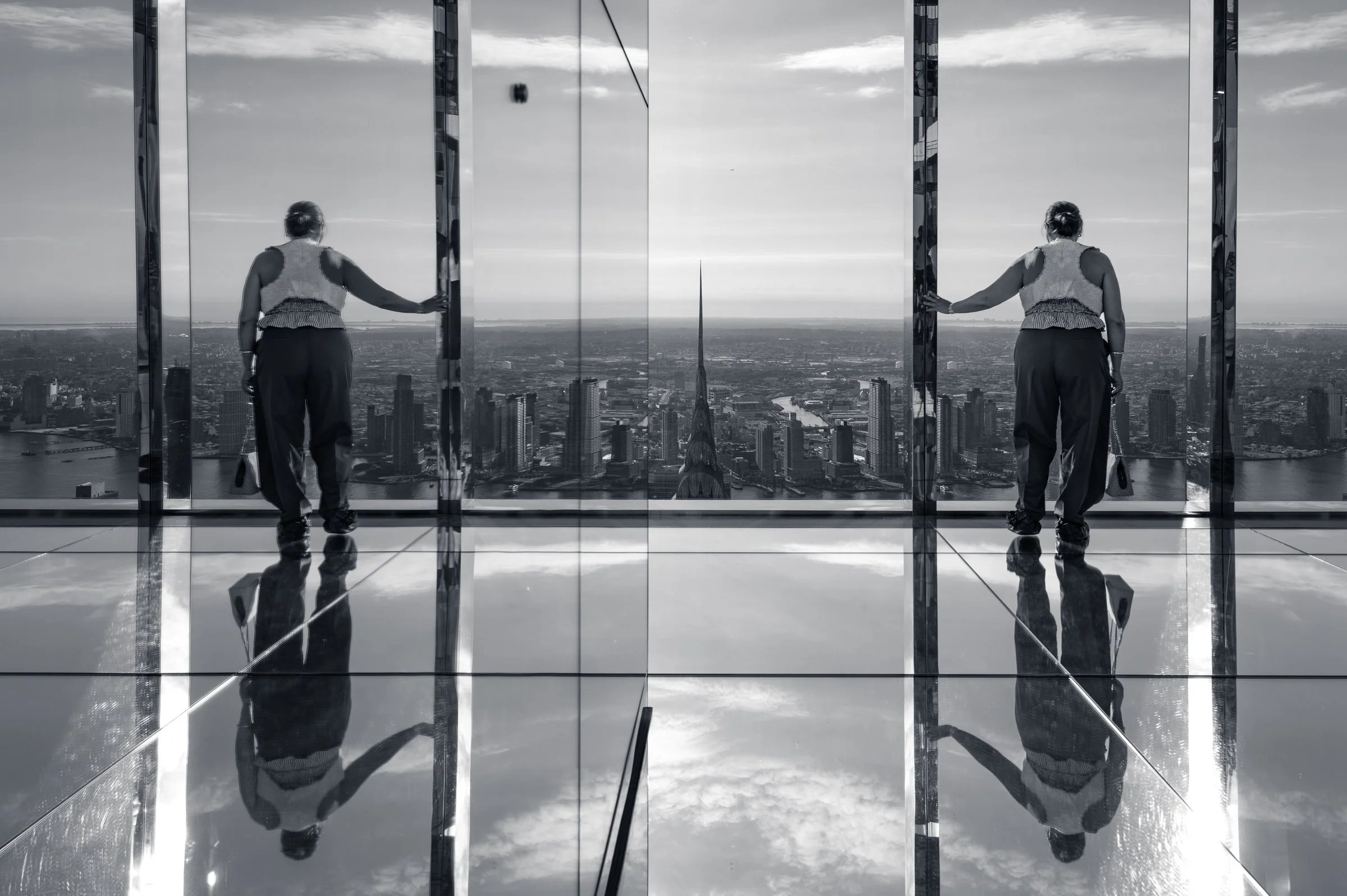 A person leaning on a glass wall doubled by the mirrored panels beside them and doubled again by the mirrored floor, a tall spire standing between the two figures, in black and white.
