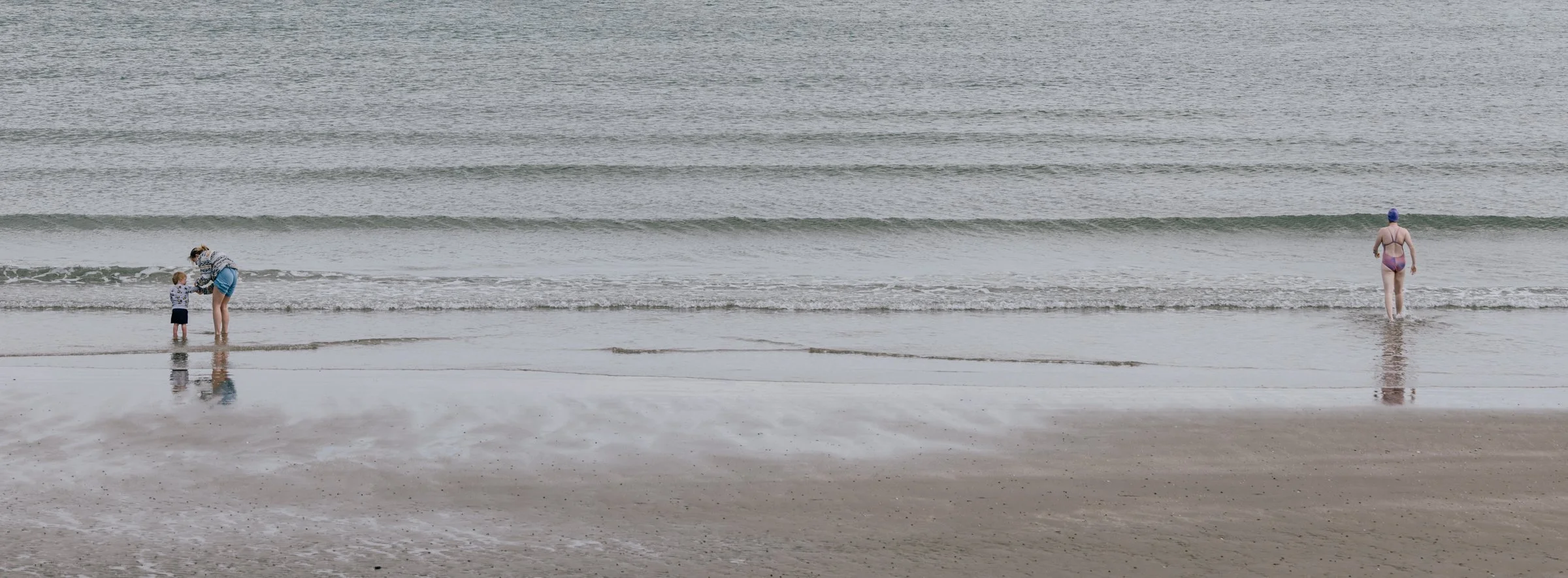 A wide pale strand at low tide, two small figures at the left edge and a single bather far off to the right, sand and water almost merging.
