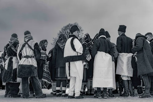 A group in traditional embroidered dress gathered close together in the snow, seen from behind, Bucovina, Romania, in black and white.