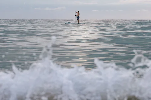 A lone paddleboarder standing on calm water beyond a breaking wave, Miami Beach.