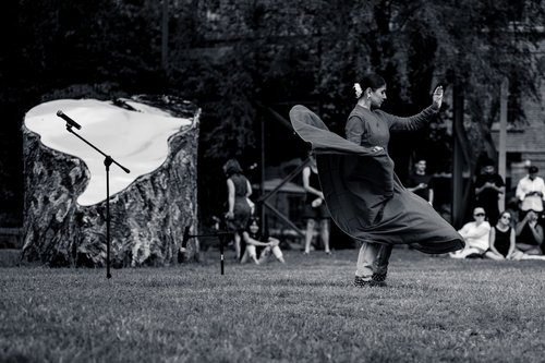 A dancer in flowing dark clothing mid-movement on the grass beside a billowing white flag, onlookers seated among the trees at Astoria Park, New York, in black and white.