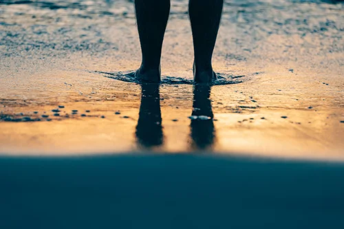 A person's legs standing in the shallow water at sunrise, reflected in the wet sand, Miami Beach.