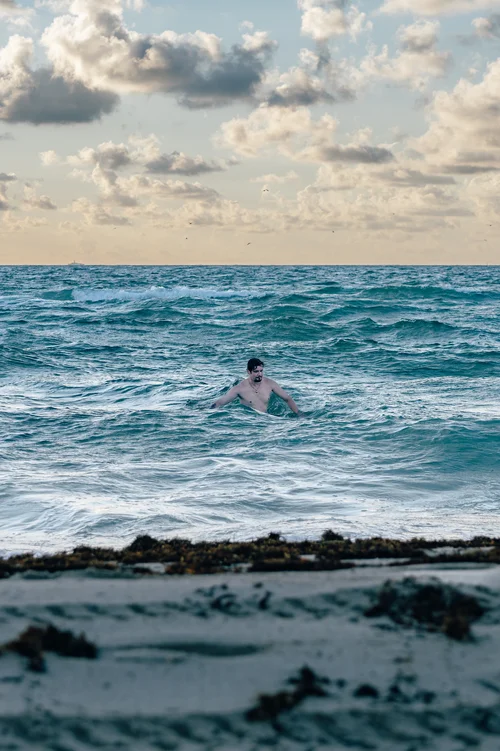 A swimmer in the surf among low rocks under a cloudy sky, Miami Beach.