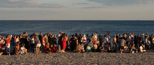 A large crowd gathered along the shoreline at the water's edge in warm dusk light at Rockaway Beach, New York.