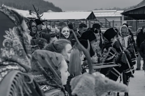 A festival crowd in elaborate costumes and feathered headdresses gathered in a village square, Bucovina, Romania, in black and white.