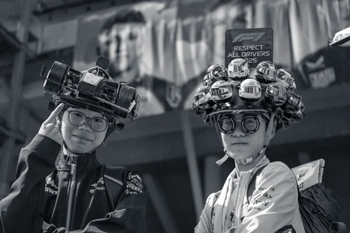 Black and white photo of two young Formula 1 fans wearing playful homemade hats decorated with model racing cars, an F1 banner behind them