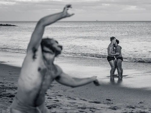 A dancer lunging with arms outstretched in the foreground while a couple stands in the shallows behind, at Rockaway Beach, New York, in black and white.