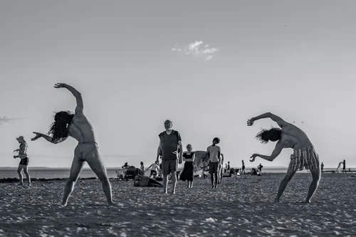 Two dancers leaping and bending across the sand, beachgoers scattered behind them under a wide sky at Rockaway Beach, New York, in black and white.
