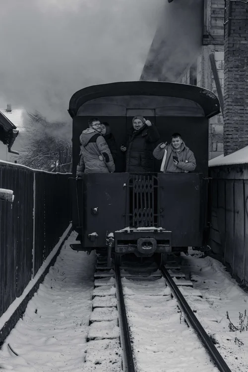 A steam locomotive on narrow-gauge tracks trailing smoke, figures gathered alongside in the snow, Bucovina, Romania, in black and white.