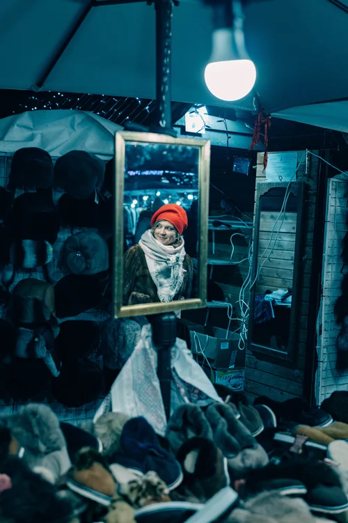 A figure in a red hat reflected in a framed mirror propped among stacked firewood at a market stall lit by a bare bulb, Bucovina, Romania.