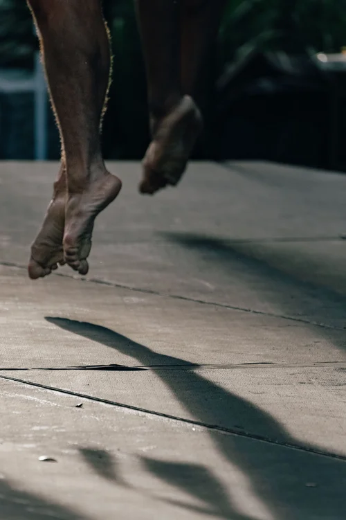A close view of a dancer's bare feet rising onto the toes on sunlit pavement, a long shadow cast beside them, Miami.