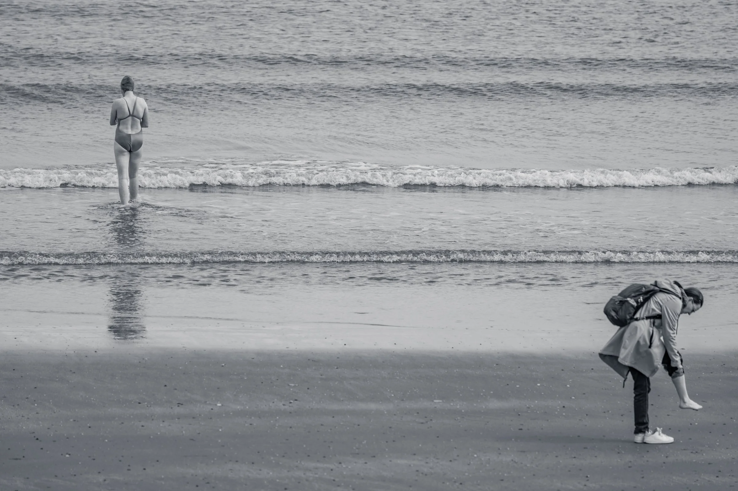 A lone figure walking out into the shallows of a wide flat sea, in black and white, low waves running across the frame.