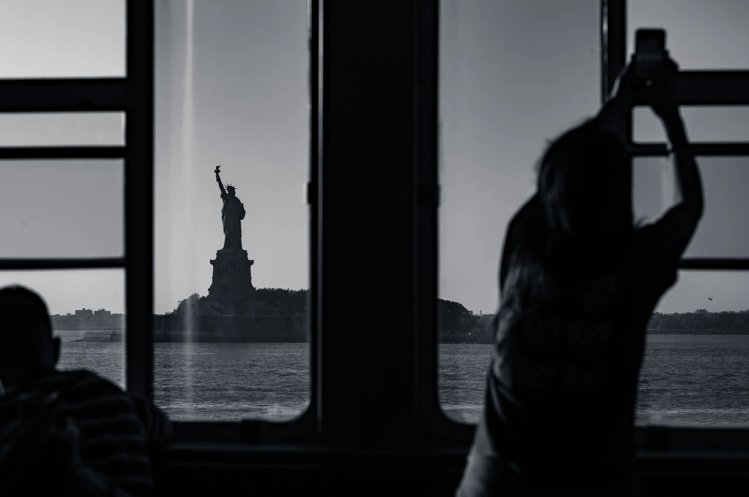 The Statue of Liberty seen at the centre through the window mullions of the Staten Island Ferry, a woman in the right foreground holding a phone up to photograph it, flat grey water beyond, in black and white.