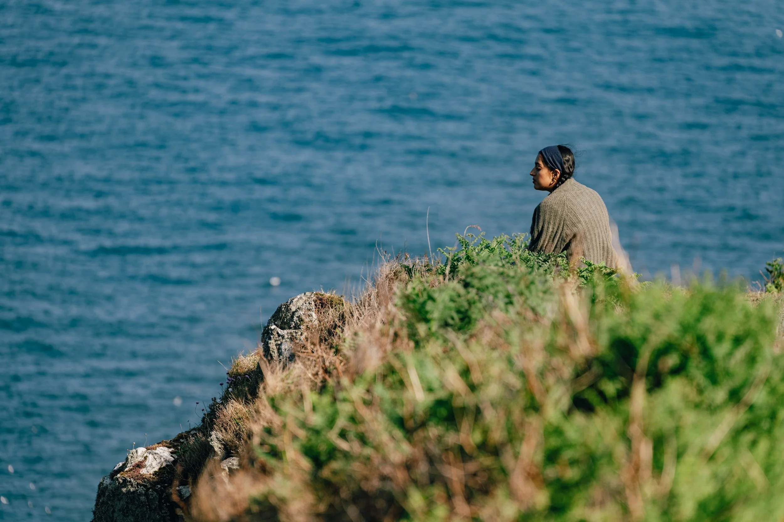 A woman sits near the cliff edge, looking out over the calm blue water, framed by green coastal foliage.