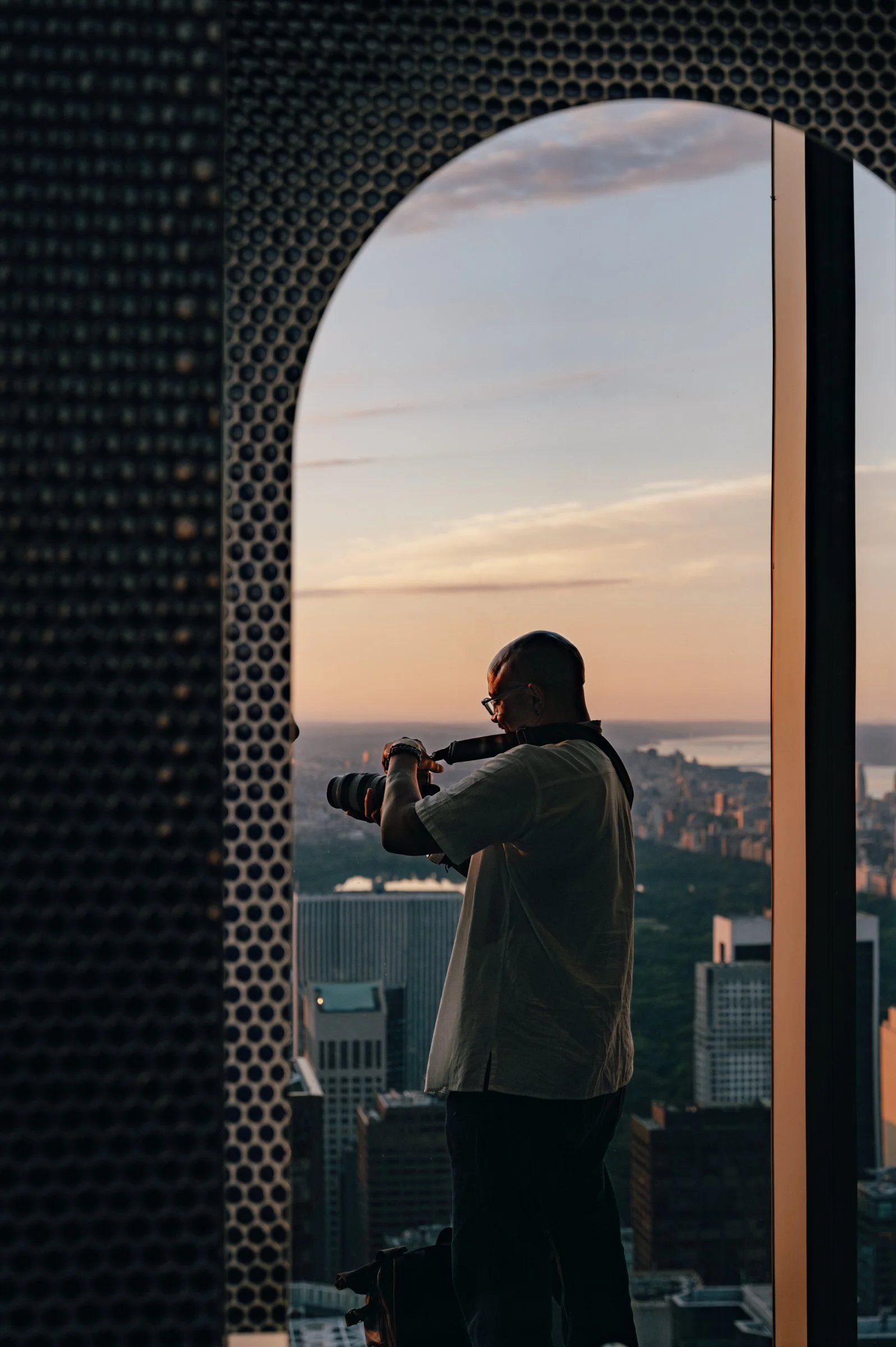 A man in a pale shirt photographing the sunrise through an arched opening in a dark perforated screen, the sky pink behind him.