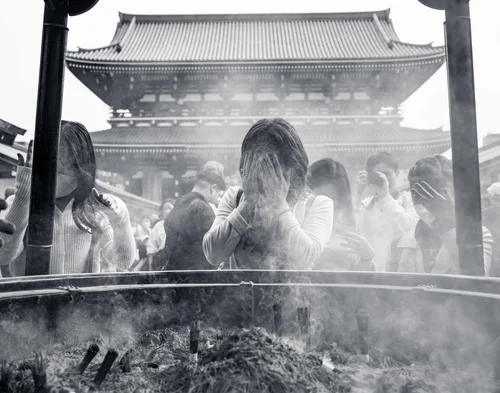 Visitors wafting incense smoke from the great cauldron before the temple hall at Senso-ji, Tokyo, in black and white.