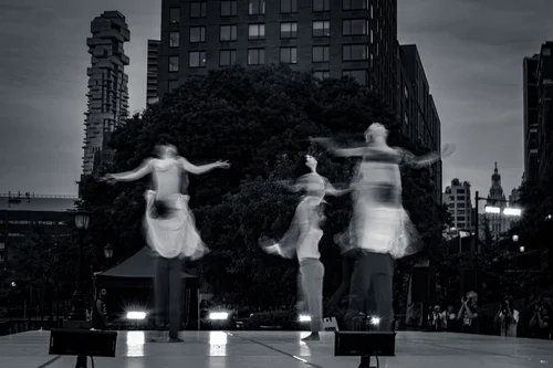 Three dancers in light clothing leaping with arms outstretched, a city building rising behind them at Battery Park, New York, in black and white.