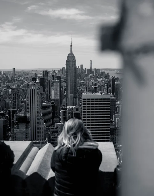 A lone person seen from behind, gazing out over the Manhattan skyline and the Empire State Building from a high window.