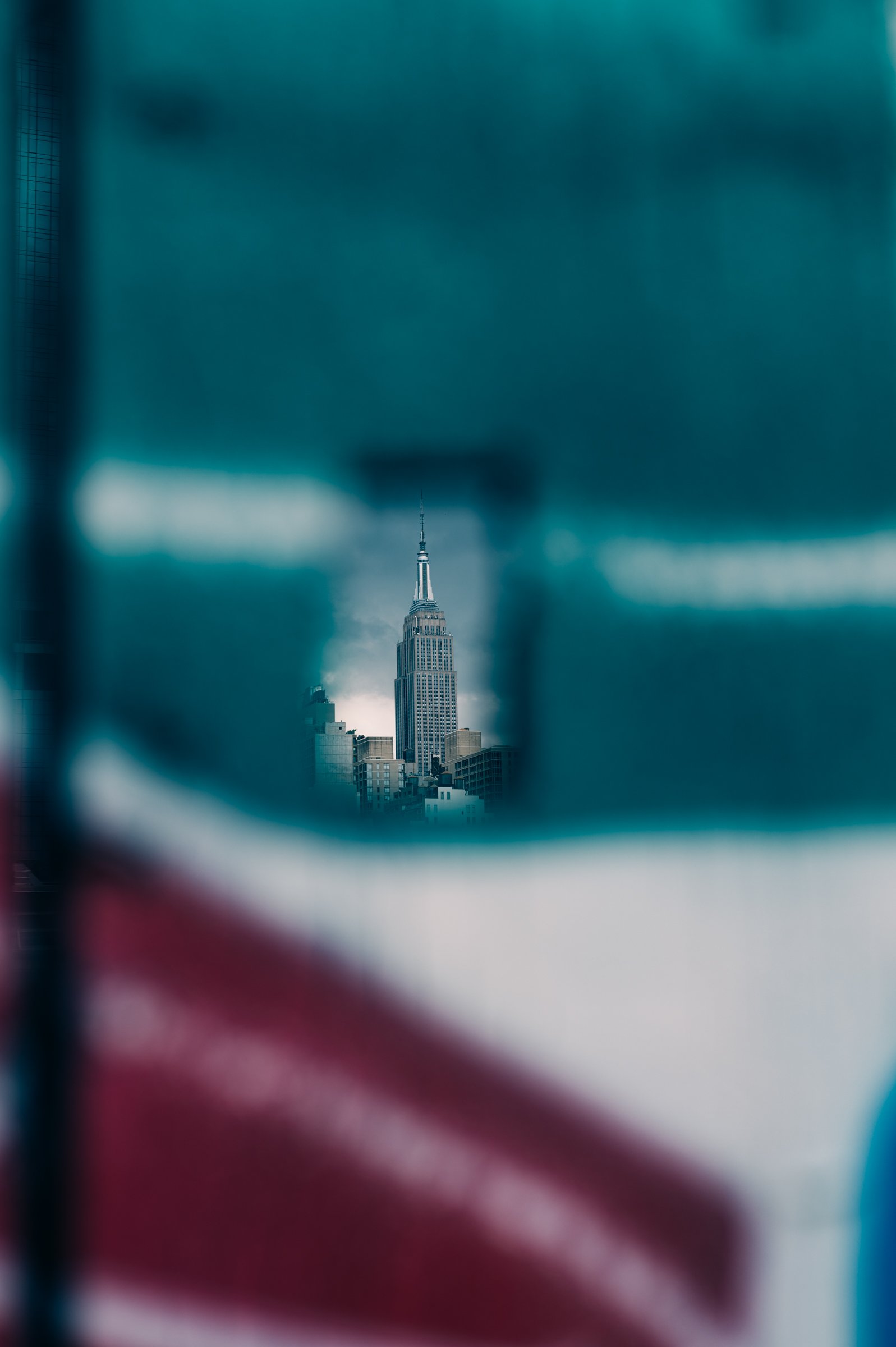 Almost the whole picture is an out-of-focus teal-green painted hoarding, with a small sharp gap in it showing the upper floors and mast of the Empire State Building.