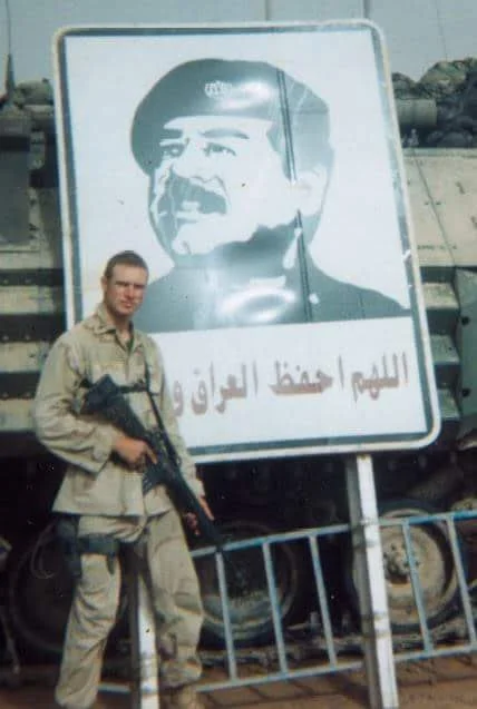 A soldier standing in front of a large sign with a portrait of a man wearing a military cap, featuring Arabic text.