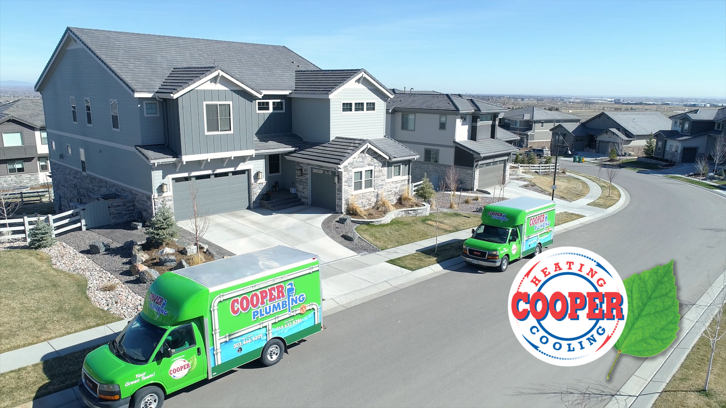 A modern, multi-story house with a gray roof and a combination of gray and white exterior walls, situated in a suburban neighborhood. Two green Cooper Plumbing service vans are parked on the driveway and street, with landscaping that includes small t
