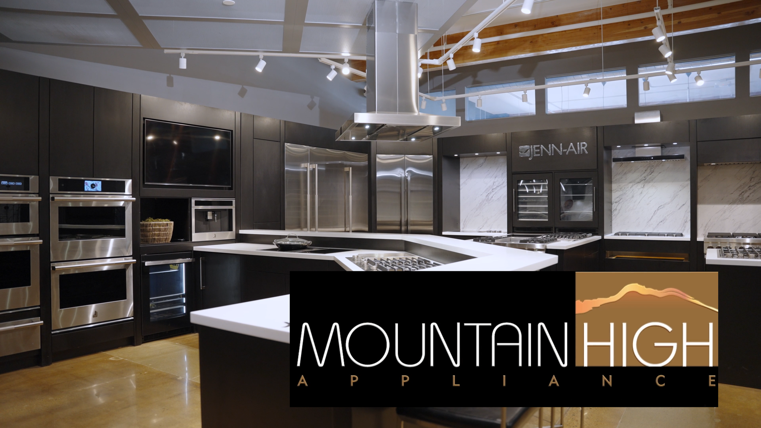 Modern kitchen with black cabinets, stainless steel appliances, a white countertop, and a stainless steel range hood. There is a large black sign with white and gold text that reads 'MOUNTAIN HIGH APPLICATION'.