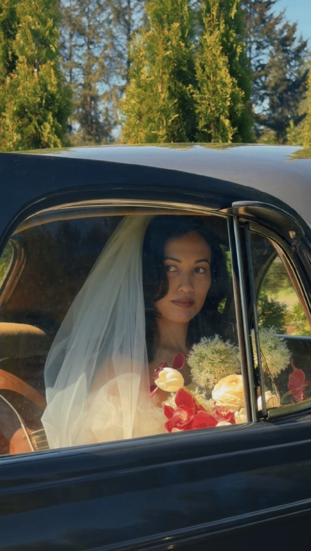 A woman in a wedding dress and veil sitting inside a vintage black car, holding a bouquet of flowers, looking outside through the window. Outside, green trees and a clear blue sky are visible.