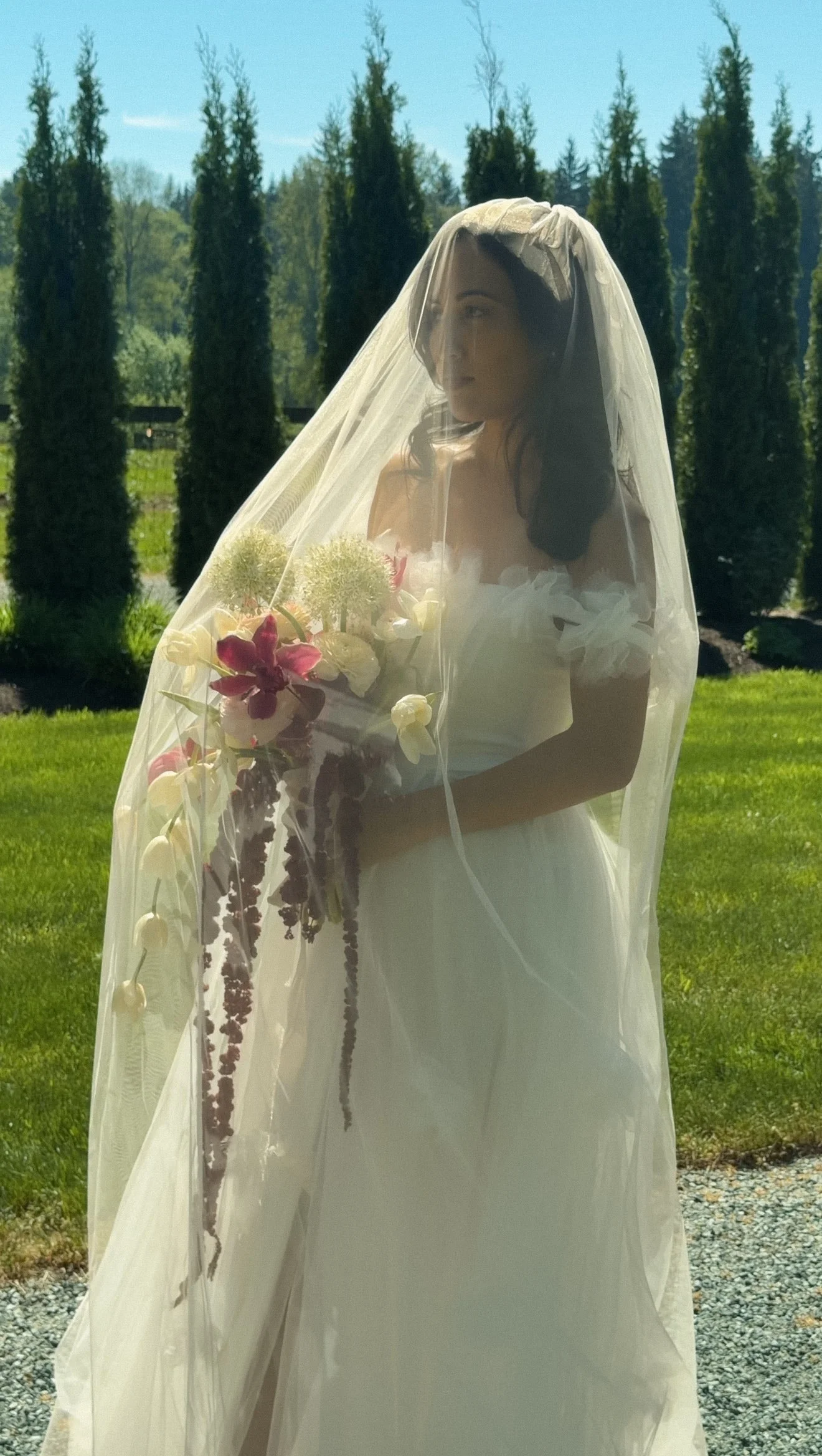 A woman in a wedding dress with a veil holding a bouquet of flowers, standing outdoors on a sunny day with a row of tall trees in the background.