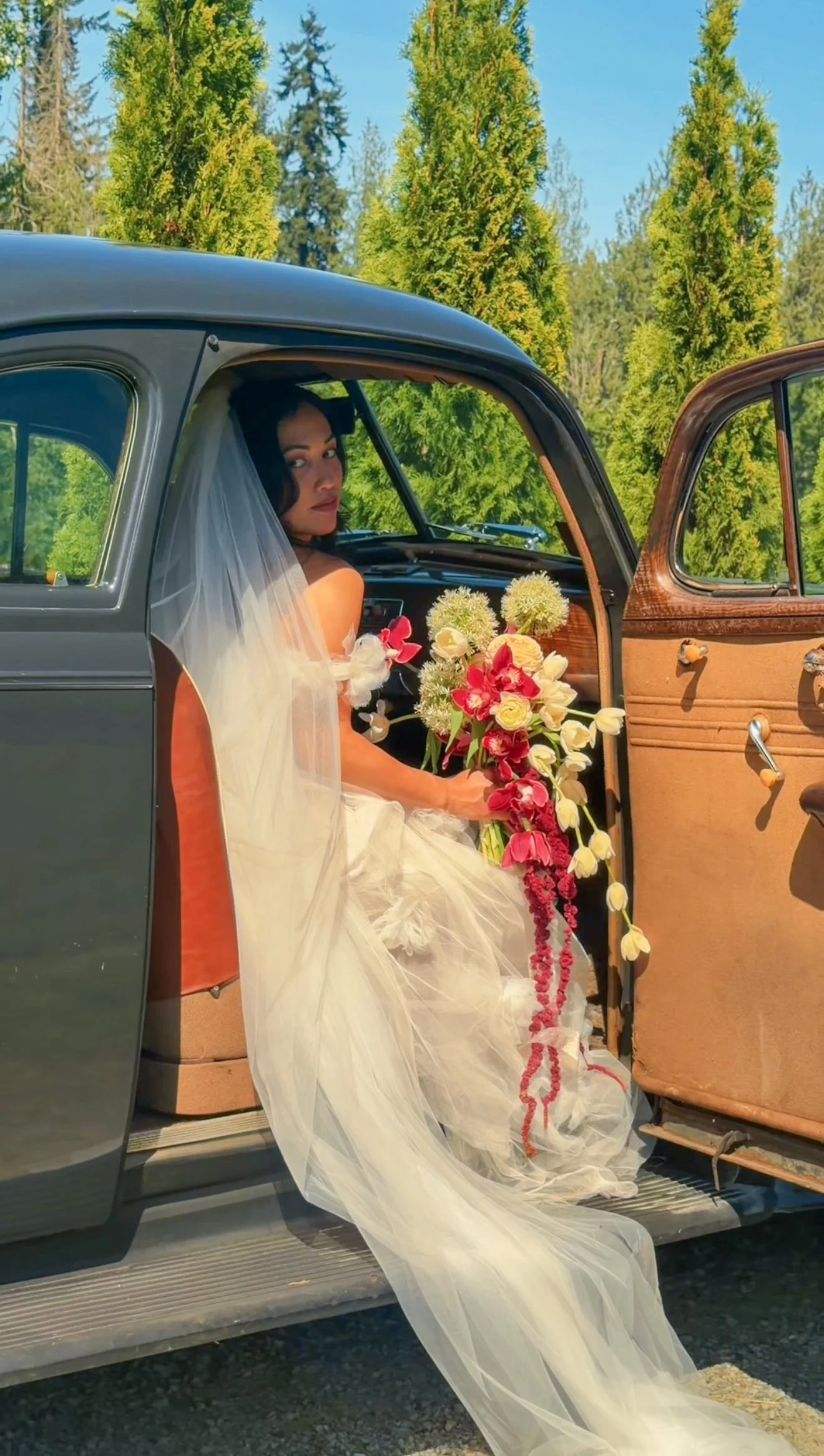 A woman wearing a wedding dress and veil is sitting inside an open vintage car, holding a bouquet of flowers. The car door is open, revealing the interior, with a backdrop of tall green trees and a clear blue sky.