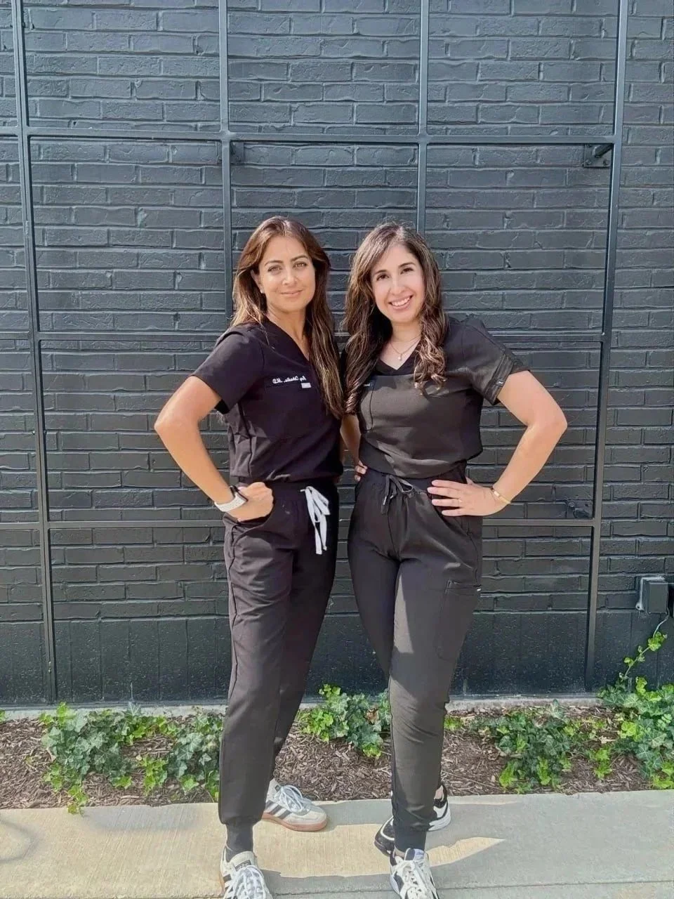 Two women standing side by side in black scrubs in front of a black brick wall and greenery, smiling at the camera.