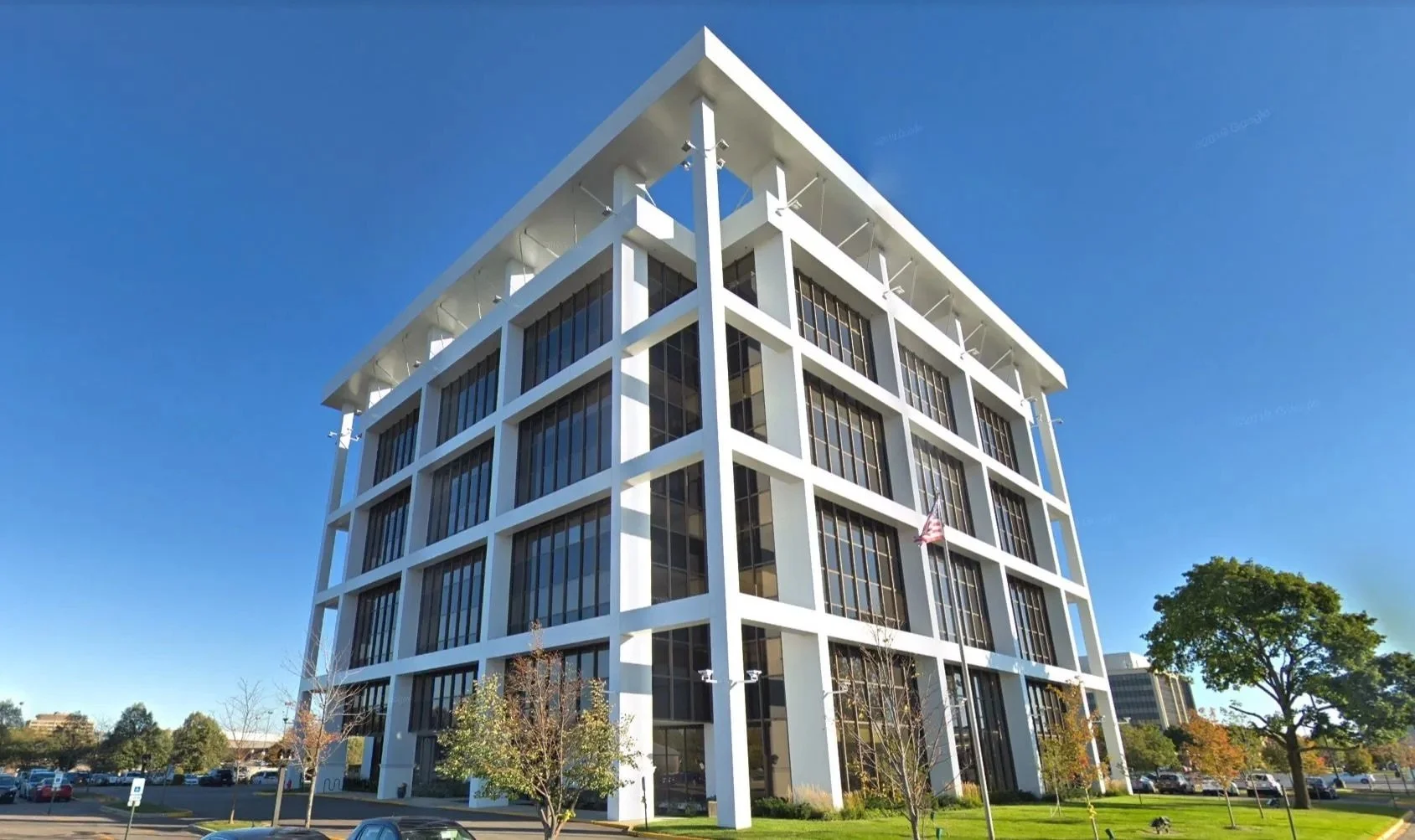 Modern multi-story office building with large glass windows, surrounded by trees and a parking lot, under a clear blue sky.