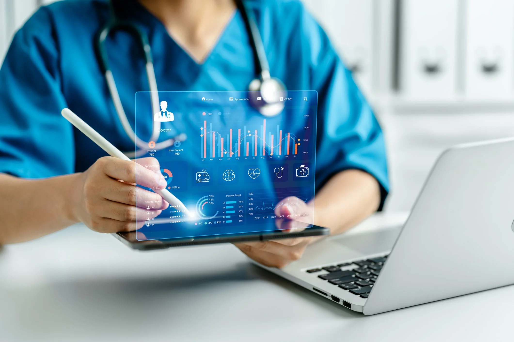 A healthcare professional in blue scrubs with a stethoscope around their neck uses a stylus to interact with a transparent digital interface displaying medical data, graphs, and icons, with a laptop on the table.