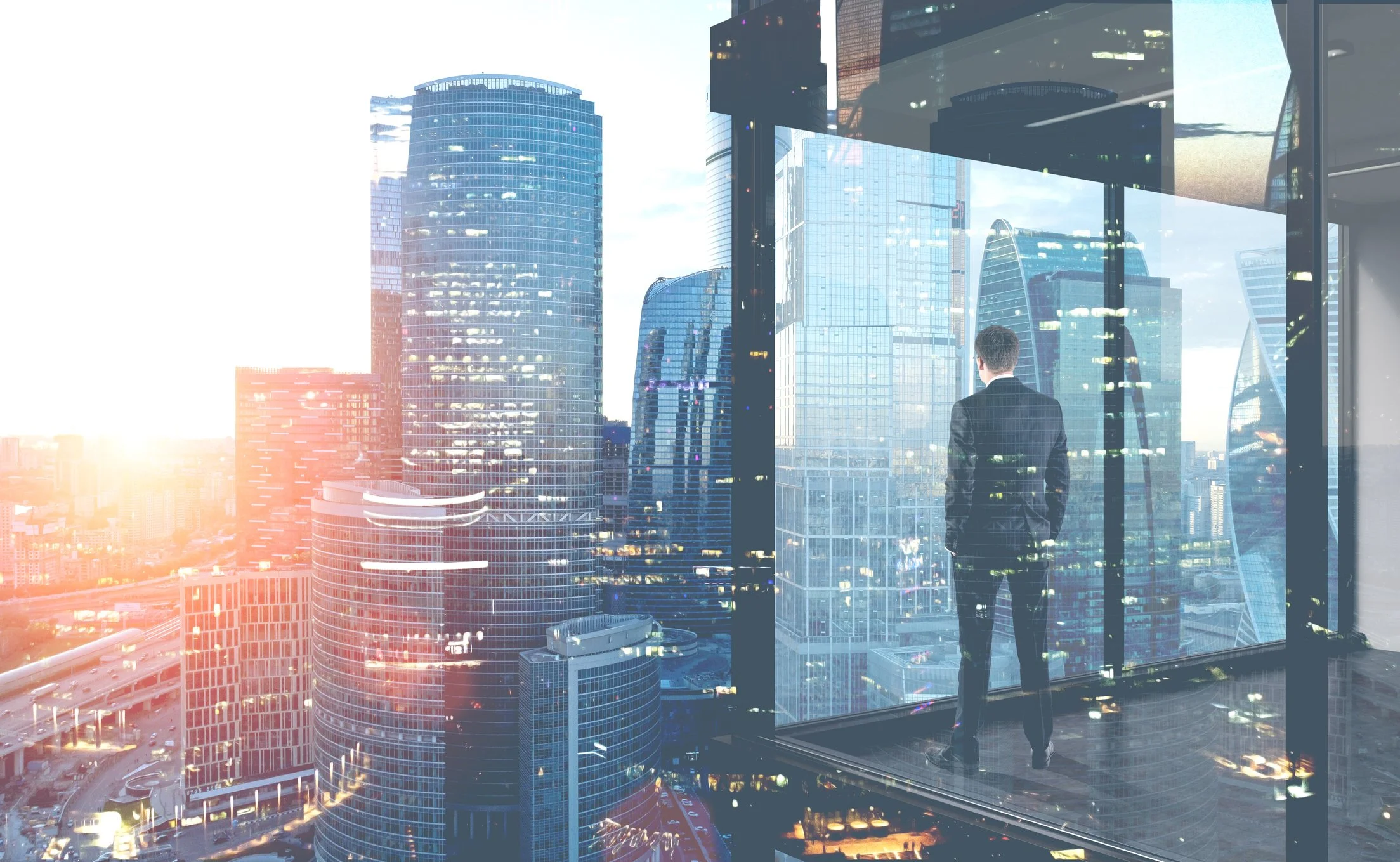 A man in a suit standing inside a skyscraper, looking out of large glass windows at a city skyline with tall modern buildings during sunset.
