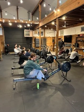 People working out on rowing machines in a gym with wooden beams and string lights.