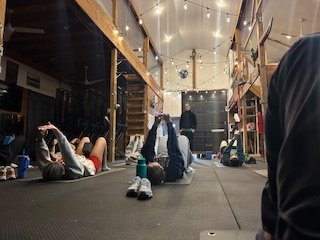 People lying on their backs on the floor inside a gym or training facility, with some sunlight and hanging lights overhead.