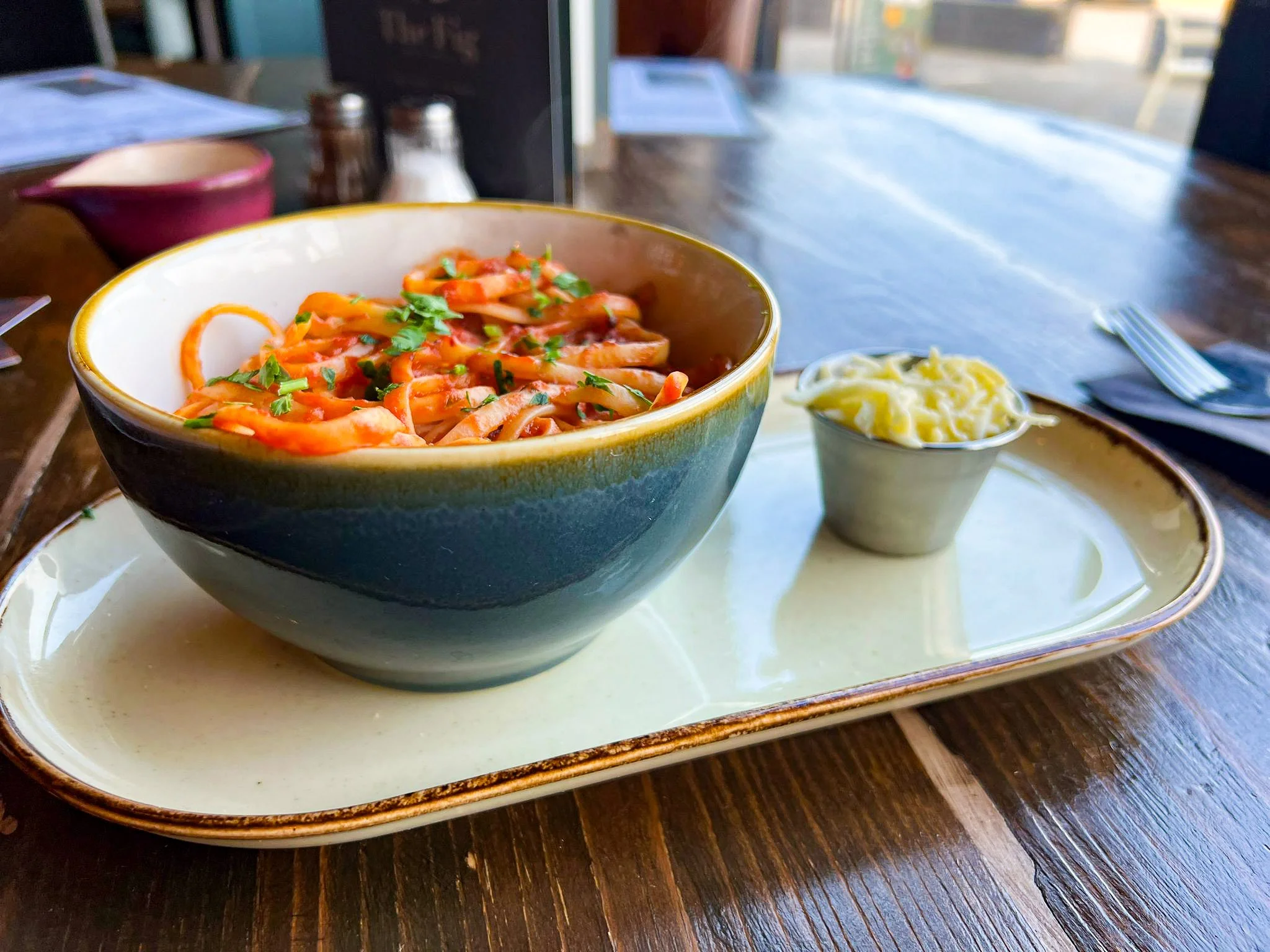 A bowl of pasta with red sauce topped with chopped herbs, served on a ceramic plate with a side of shredded cheese in a small cup, all on a wooden table in a restaurant.