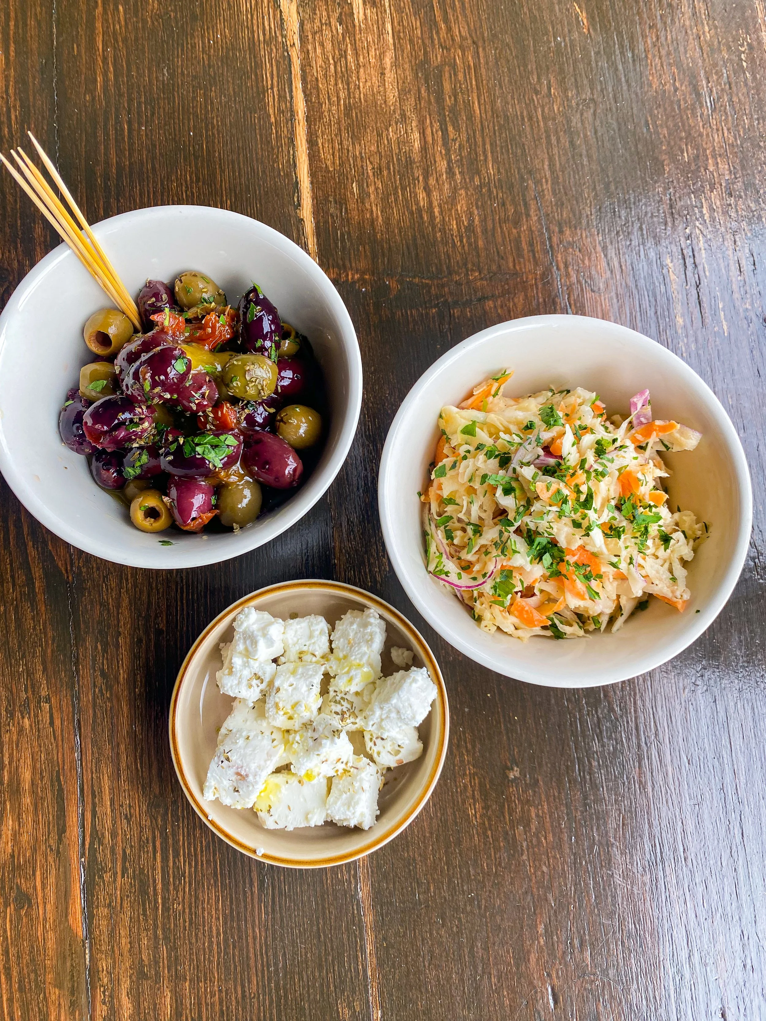 A bowl of green and purple olives garnished with herbs, a bowl of coleslaw with shredded carrots, cabbage and herbs, and a bowl of cubed feta cheese on a wooden table.