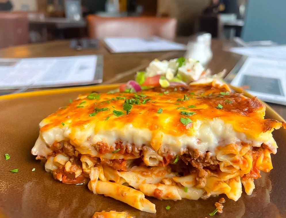 Close-up of a serving of baked lasagna on a brown plate, topped with melted cheese and garnished with chopped parsley, with a side of salad and a shaker of grated cheese in a restaurant setting.