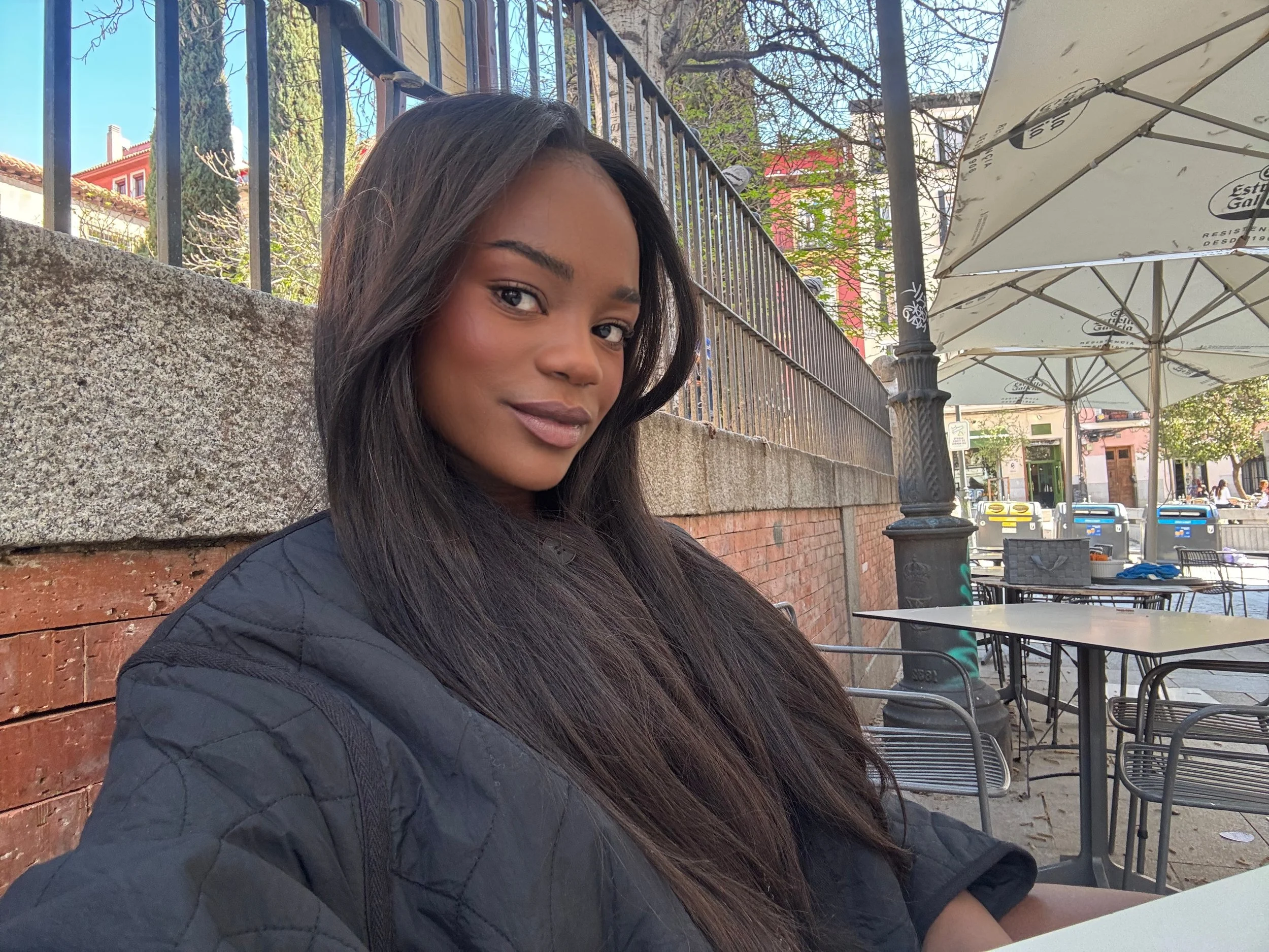 Young woman with long dark hair and a soft smile sitting at an outdoor café on a sunny day, with tables, chairs, and umbrellas visible in the background.