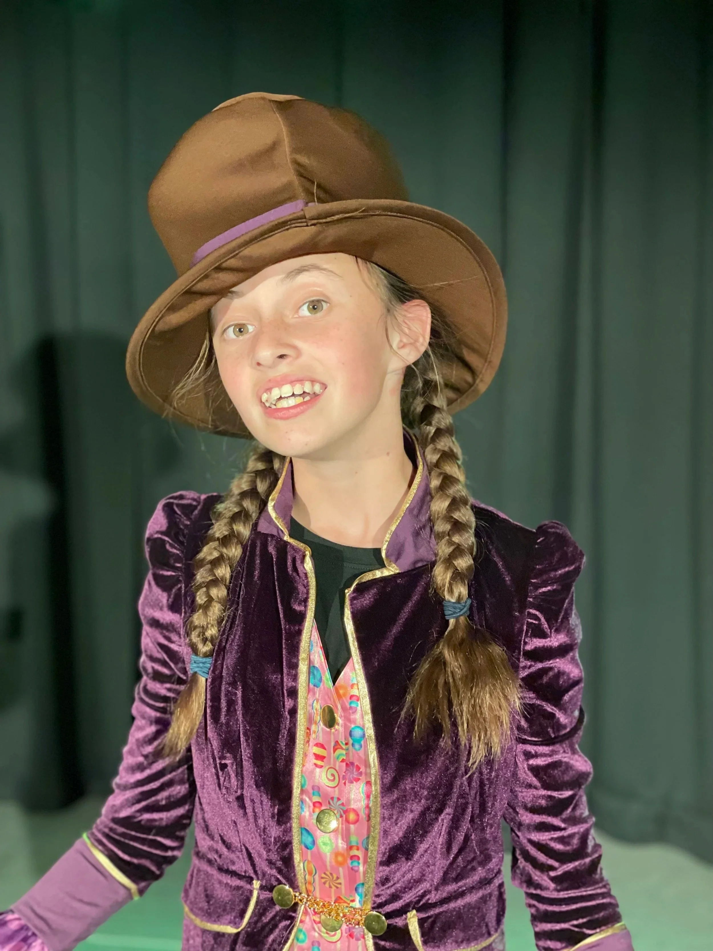 Girl dressed as a witch, wearing a large brown hat, purple velvet jacket, and a colorful dress with candy patterns, posing in front of a dark backdrop.