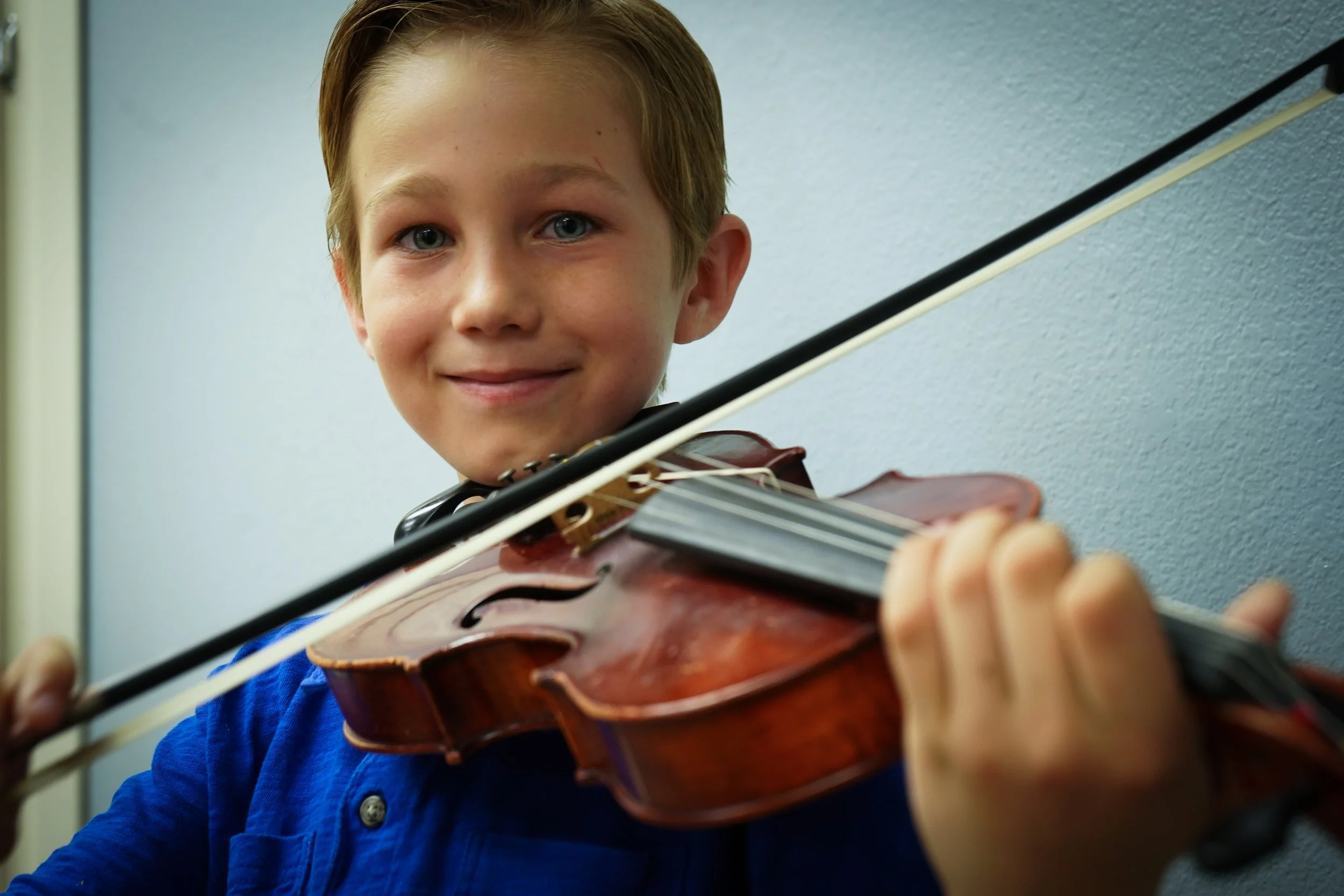 Young boy playing violin and smiling, wearing a blue shirt, indoors against a plain wall.