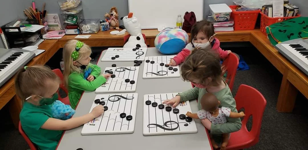 Five children seated at a table with musical note boards and black counters, engaging in a musical activity. One child has a toy baby doll, and another is holding a stuffed animal. In the background, shelves with books, toys, and a keyboard are visib