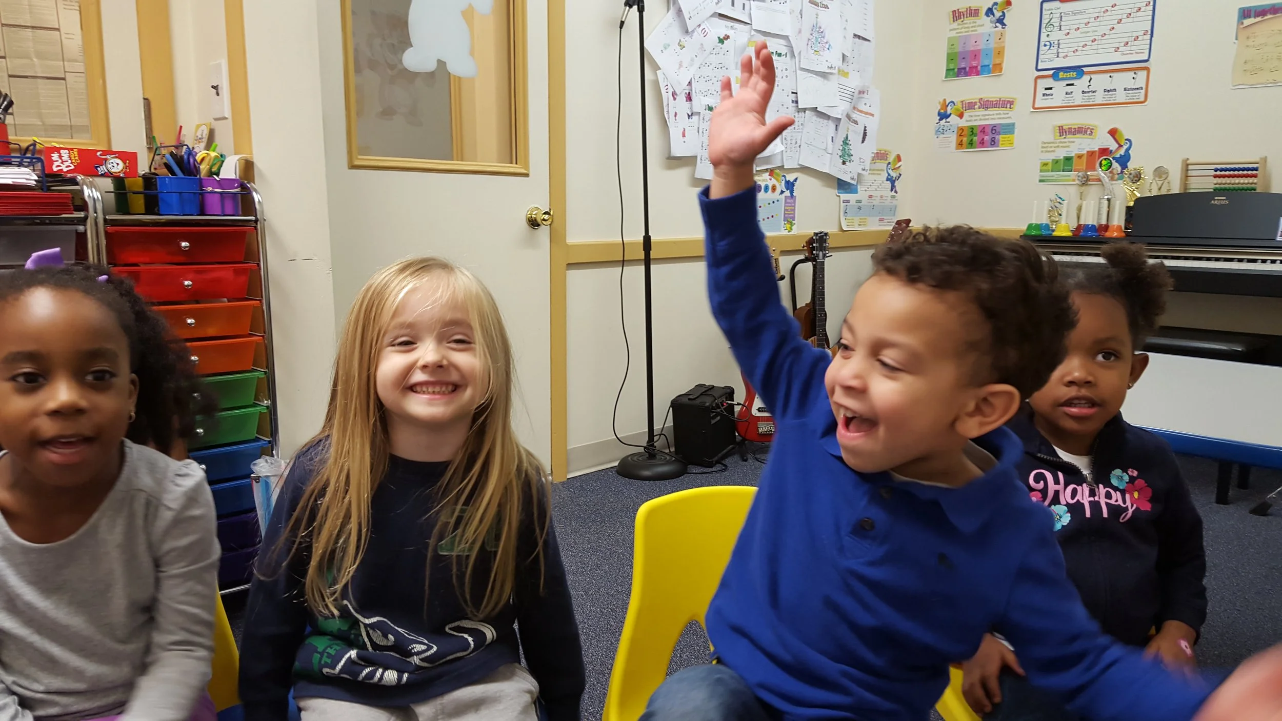 Four children sitting in a classroom, smiling and engaging. A boy with curly hair wearing a blue shirt has his hand raised, and the other children are watching him. The classroom has colorful posters, a musical keyboard, and a cart with colorful supp