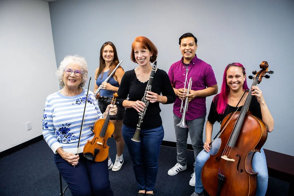Five women holding musical instruments, including a violin, clarinet, trumpet, and cello, standing together in an indoor setting with plain walls, smiling at the camera.